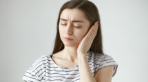 Studio shot of attractive casually dressed young dark haired female keeping eyes closed and covering sore ear with hand while having some problems with hearing. Health, people and pain concept