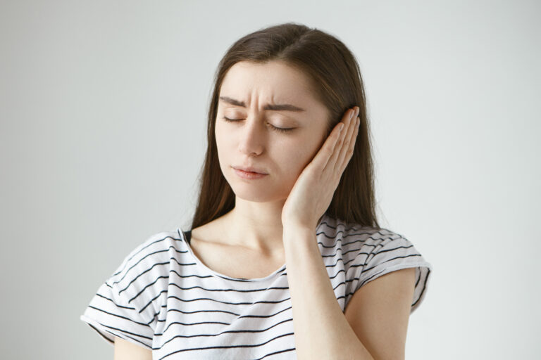 Studio shot of attractive casually dressed young dark haired female keeping eyes closed and covering sore ear with hand while having some problems with hearing. Health, people and pain concept